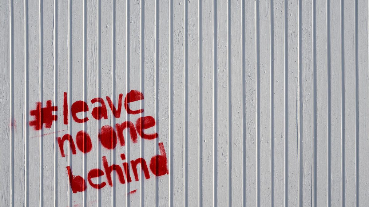 The hashtag Leave No One Behind is spraypainted on a white wall in red paint.